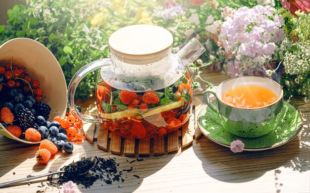 Thé aux fruits aromatiques naturels dans une théière transparente et dans une tasse parmi les baies et les fleurs. thé aromatique réchauffant avec un arôme profond de baies et de fleurs sauvages.