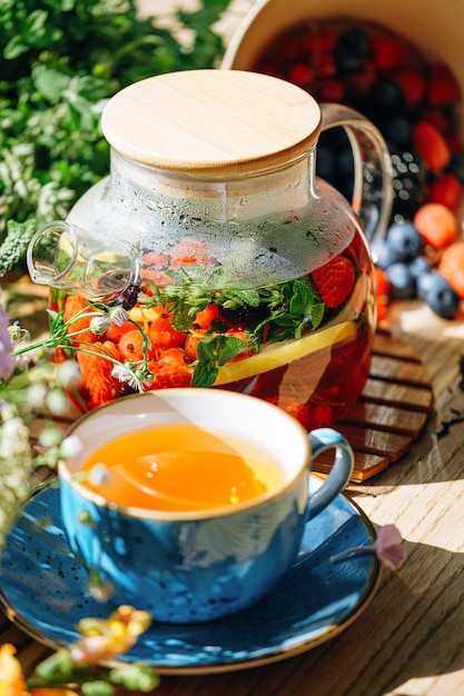 Thé aux fruits aromatiques naturels dans une théière transparente et dans une tasse parmi les baies et les fleurs thé aromatique réchauffant avec un arôme profond de baies et de fleurs sauvages