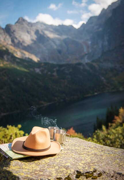 Thé chaud dans une tasse touristique en métal et un chapeau sur fond de beau paysage de bois et de montagnes.