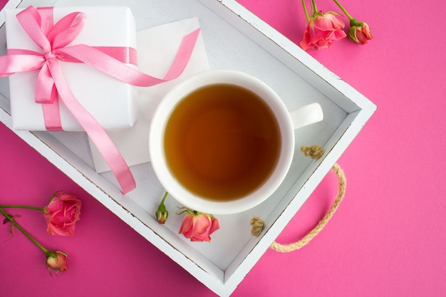 Thé dans la tasse et cadeau sur le plateau en bois blanc sur fond rose. Vue de dessus.