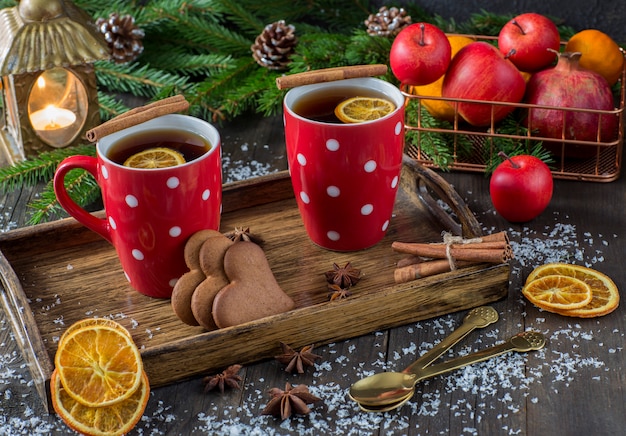 Thé dans une tasse avec un décor tricoté rouge, une lanterne, des tranches d'orange, une corbeille de fruits, de la cannelle, des branches d'épinette et des biscuits en forme de cœur