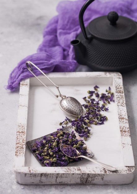 Thé de fleurs de mauve bleue en plaque d'acier avec infuseur à passoire vintage dans une boîte en bois avec théière et tissu violet sur fond blanc