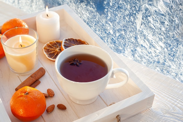 Thé d'hiver à la fenêtre givrée. Thé aux mandarines, cannelle et noix dans une tasse blanche sur un plateau en bois blanc. Photo de haute qualité