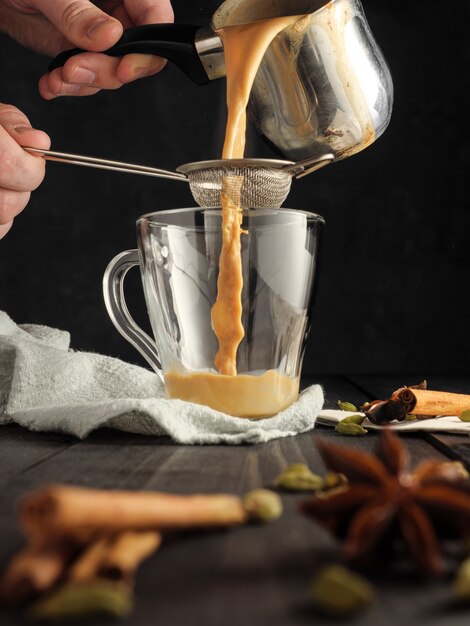 Le thé Masala est versé à travers un tamis dans une tasse en verre. Les espèces sont sur la table.