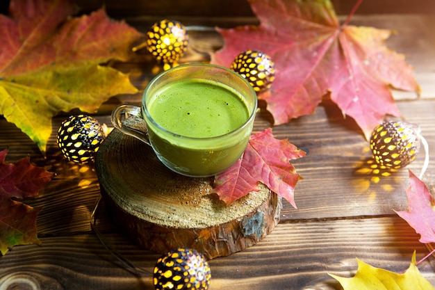 Thé matcha japonais vert avec de la mousse dans une tasse transparente sur une table en bois en automne nature morte. Ambiance chaleureuse et confort, guirlandes lumineuses, feuilles d'érable rouge, bâtons de cannelle, citrouille, biscuits, tranche.