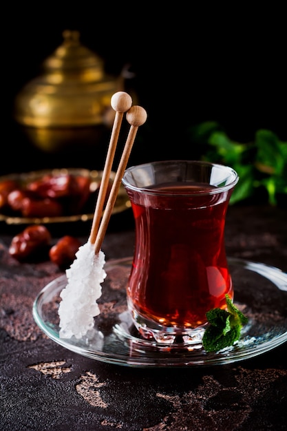 Thé turc dans une tasse en verre traditionnelle avec du sucre caramélisé et de la menthe sur une table sombre.