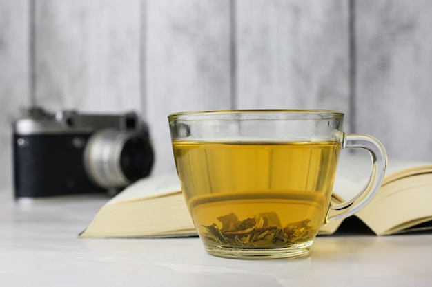 Thé vert aux herbes infusé dans une tasse transparente à côté d'un livre ouvert et d'un appareil photo vintage