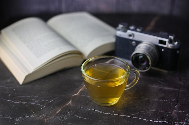 Thé vert aux herbes infusé dans une tasse transparente près d'un livre ouvert et d'un appareil photo vintage sur fond sombre