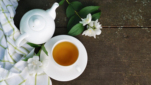 Thé vert chaud dans une théière et une tasse avec une branche de fleurs de jasmin et une serviette blanche sur fond de bois brun rustique rugueux. Vue de dessus, concept de vie lente et saine de la nature.