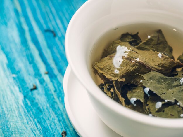 Le thé vert est infusé dans un bol blanc sur un fond en bois bleu. Des feuilles de thé sèches se trouvent à proximité, gros plan