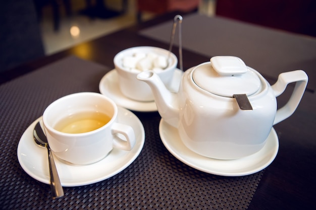 Théière blanche avec tasse à thé, soucoupe et cuillère à café, un sucrier sont sur la table