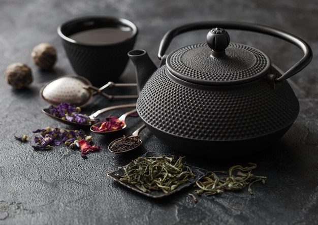 Théière chinoise avec tasse et infuseur avec divers thés sur fond noir Bourgeons de rose fleurs de mauve bleue thé en vrac vert et noir
