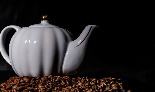 Théière en porcelaine parmi les grains de café sur fond noir.