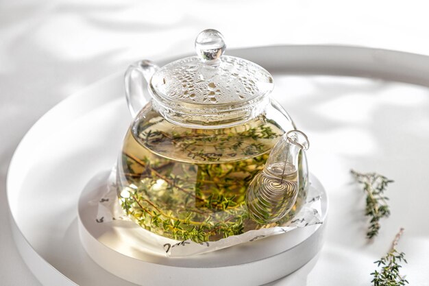 Théière de tisane chaude au thym sur plateau blanc et fond blanc sous les rayons du soleil