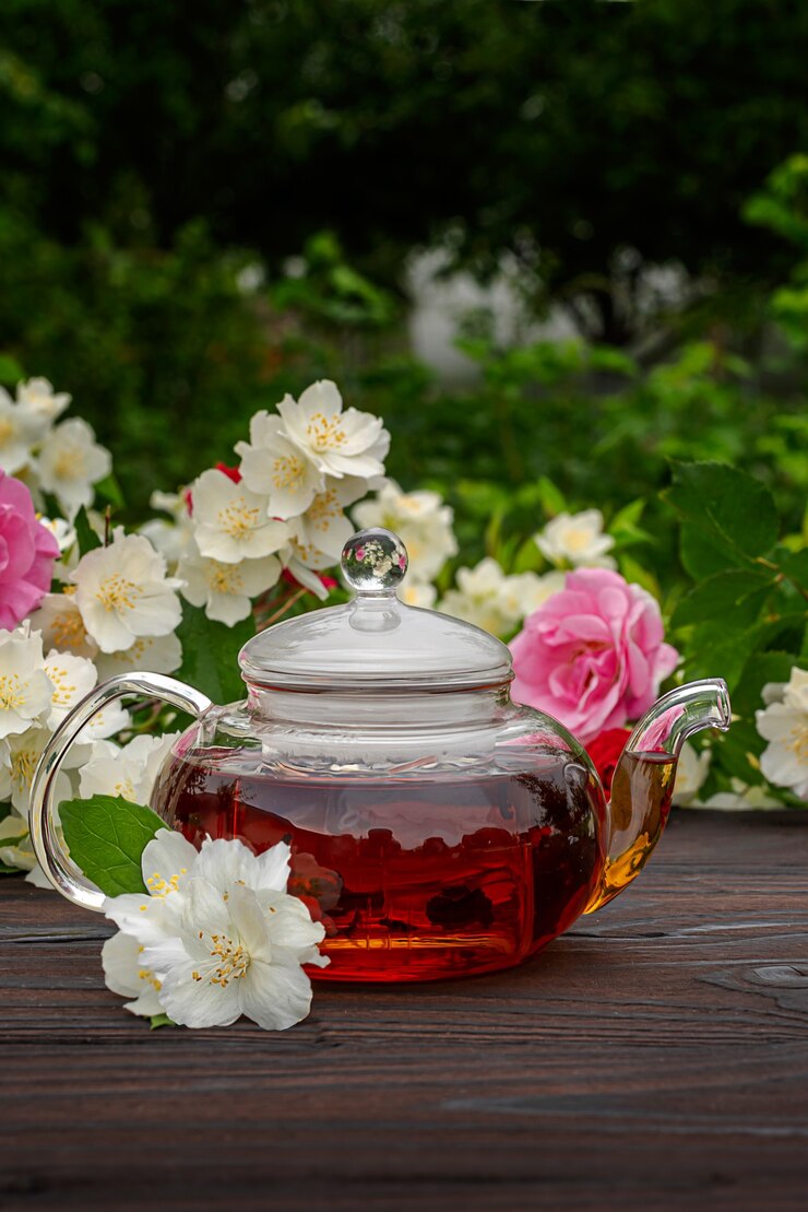 Théière en verre cuite à la vapeur sur une table en bois parmi des buissons fleuris de roses et de jasmin