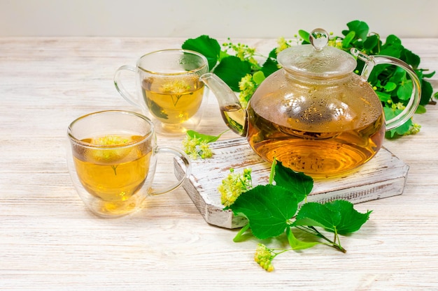 Une théière en verre et deux tasses de thé avec un tilleul sur une table en bois sur fond de feuilles et de fleurs de tilleul