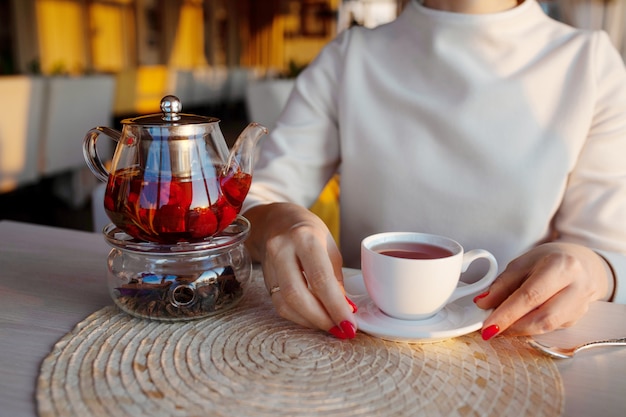 Théière en verre avec du thé au café. Mains de femme avec une tasse de thé sur fond