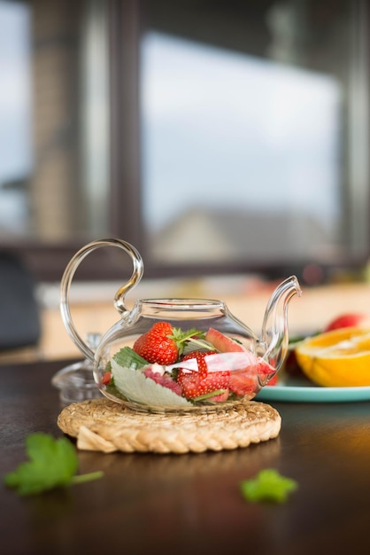 Une théière en verre avec des fruits et des baies de menthe laisse une bouteille d'eau sur la table à l'extérieur du thé aux fruits d'été