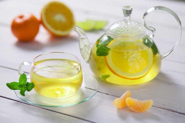 Théière en verre et tasse à thé vert, orange, mandarine, citron vert menthe sur table en bois blanc