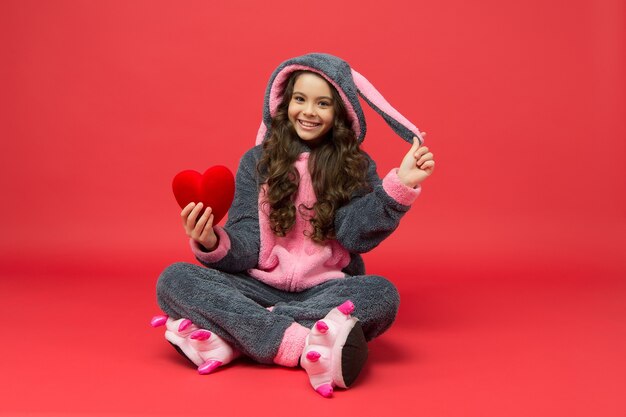 Thérapie et amour. Un enfant heureux en pyjama de lapin tient un coeur rouge. Diagnostic cardio. Chirurgie cardiaque. Maladie cardiovasculaire. Activité cardiaque. Cardiologie pédiatrique. Soins de santé. Soins de santé.