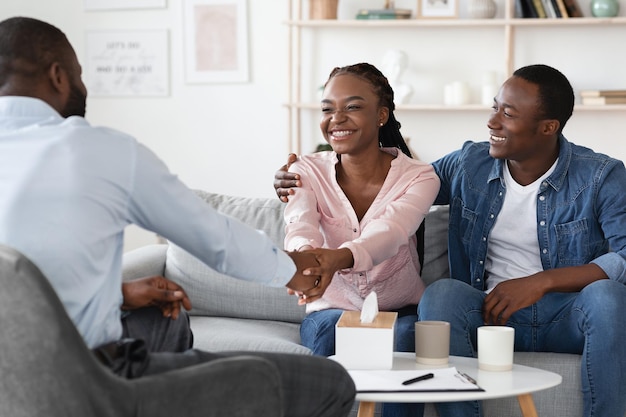 Thérapie conjugale réussie. Heureux époux noirs se serrant la main avec un conseiller familial après la réconciliation, reconnaissants pour l'aide professionnelle et le mariage sauvé, assis sur un canapé au bureau, espace libre