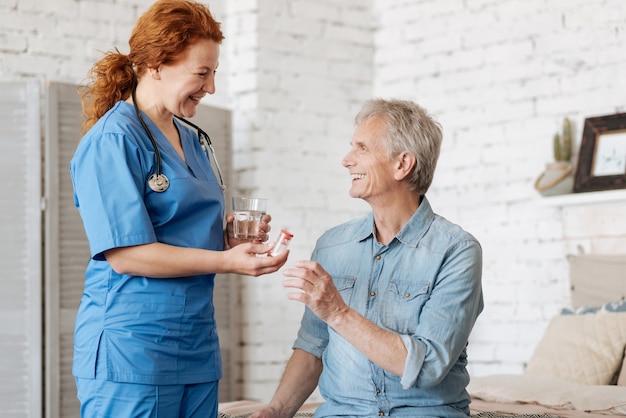 Thérapie à domicile. Neat belle infirmière proéminente servant à son patient un verre d'eau et des médicaments pour se remettre de la maladie qu'il souffre