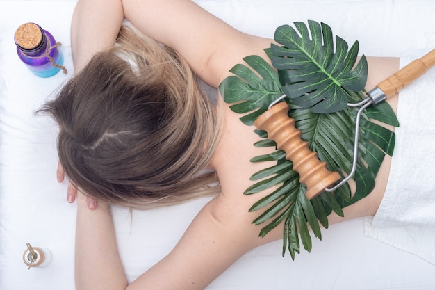 Thérapie Madero, massage relaxant - Masseur à rouleaux allongé sur des feuilles vertes sur le dos d'une femme et bouteilles d'huiles essentielles, gros plan, vue de dessus. Concept de soins du corps