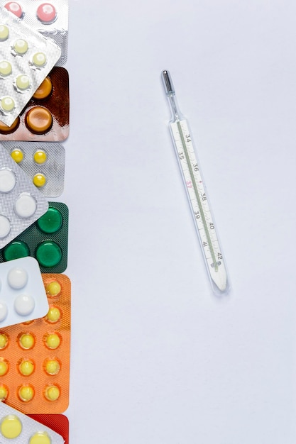 Thermomètre sur fond blanc parmi des pilules de différentes couleurs dans des emballages