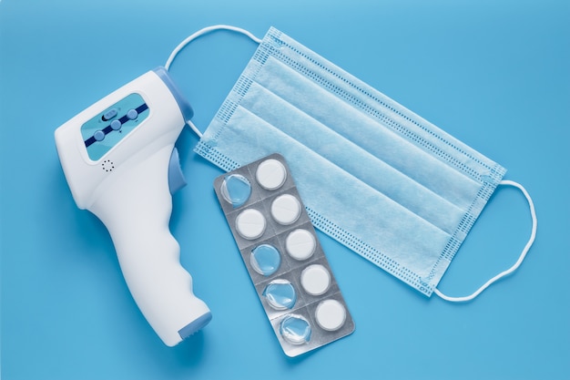 Thermomètre médical frontal infrarouge médical sans contact pour les tests de coronavirus (COVID-19). Masque chirurgical de protection et pilules blanches sur bleu