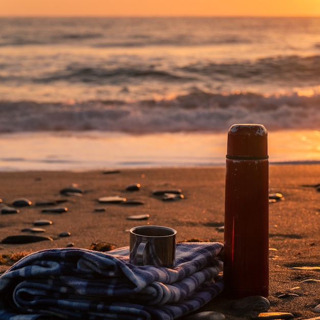 Thermos de thé chaud et un plaid doux sur une plage de sable tôt le matin