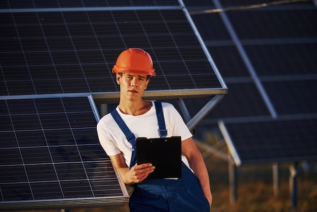 Tient le bloc-notes en main. Travailleur masculin en uniforme bleu à l'extérieur avec des batteries solaires aux beaux jours.