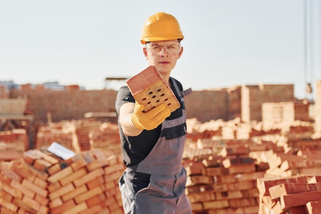 Tient la brique à la main Un ouvrier du bâtiment en uniforme et équipement de sécurité a un travail sur la construction