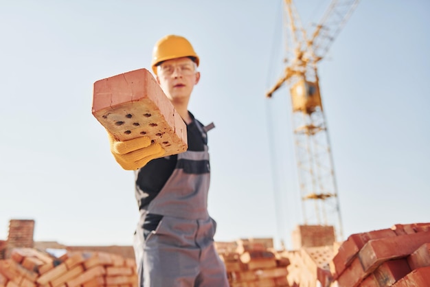Tient la brique à la main Un ouvrier du bâtiment en uniforme et équipement de sécurité a un travail sur la construction