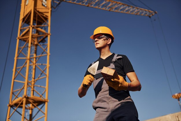 Tient la brique et l'outil dans les mains Un ouvrier du bâtiment en uniforme et équipement de sécurité a un travail sur la construction