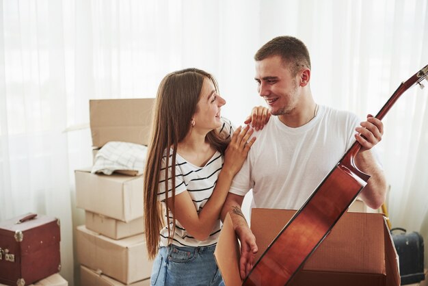 Tient la guitare acoustique dans les mains. Joyeux jeune couple dans leur nouvel appartement. Conception du déménagement.