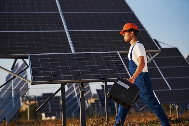 Tient la mallette avec l'équipement en main. Travailleur masculin en uniforme bleu à l'extérieur avec des batteries solaires aux beaux jours.