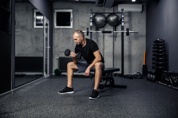 Tir de l'arrière d'un homme d'âge moyen tenant et soulevant un haltère d'une main alors qu'il était assis sur un banc de sport salle de sport moderne à l'intérieur