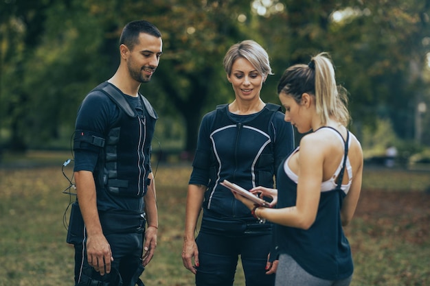 Tir d'un entraîneur personnel féminin enseignant à un couple souriant et confiant de faire des exercices avec un appareil ems dans le parc de la ville.