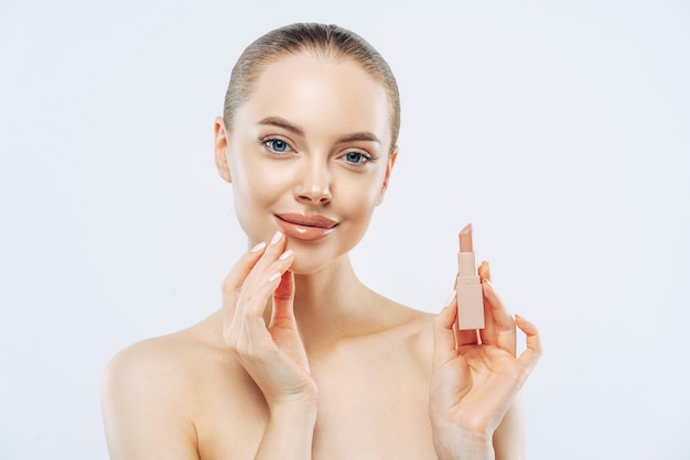 Tir isolé de la belle fille applique le rouge à lèvres sur les lèvres, a un maquillage parfait, pose avec un produit de beauté, touche doucement le visage, pose torse nu, se prépare pour le travail, pose sur fond blanc