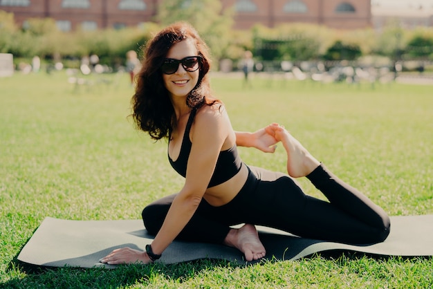 Tir en plein air d'une femme souriante sportive étend les jambes sur des sourires de karemat porte positivement des vêtements de sport a un entraînement de remise en forme pour la flexibilité fait des exercices d'étirement apprécie l'air frais et la pelouse verte.