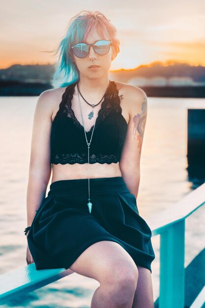 Tir vertical d'une jeune femme avec des lunettes de soleil et des tatouages dans une tenue noire sur une jetée au bord de la mer