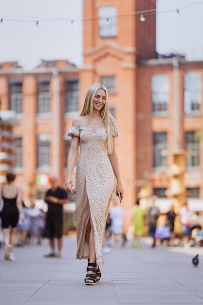 Tir vertical d'une jeune femme suédoise blonde marchant en robe beige marchant dans la rue au-dessus d'une ville floue sur fond Jolie étudiante suédoise souriante Femme reconnaissante le week-end Été de la jeunesse