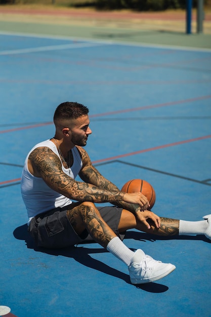 Tir vertical d'un jeune homme caucasien avec des tatouages assis sur le terrain de basket avec un ballon