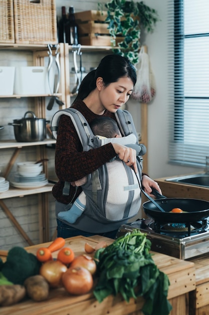 Tir Vertical Mère Asiatique Cuisine Des Aliments Avec Une Spatule Et Une Poêle à Frire Près De La Cuisinière Tout En Portant Son Nouveau-né Dans La Fronde à La Maison Pendant La Journée.