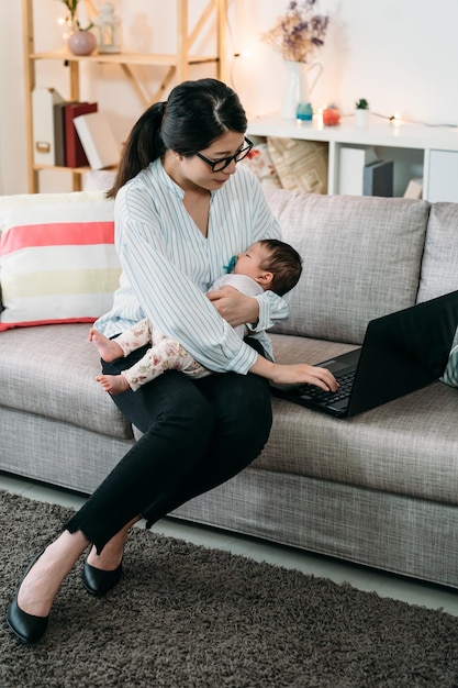 tir vertical occupé mère asiatique qui travaille tient son bébé endormi tout en utilisant l'ordinateur sur le canapé dans le salon à la maison.