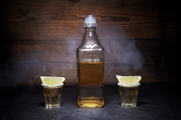 Tirs de tequila or avec des fruits de citron vert sur bois. Bouteille de tequila. Fumée.