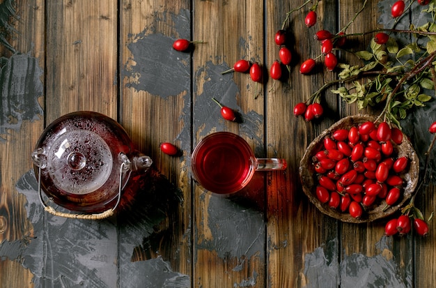 Tisane de baies d'églantier dans une théière en verre et une tasse debout sur un vieux fond de planche de bois avec des baies d'automne sauvages autour. Boisson chaude d'hiver. Mise à plat, espace de copie