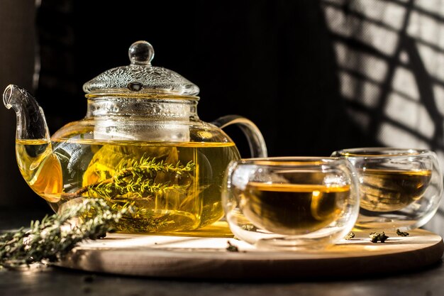 Tisane jaune dans une théière en verre transparent et deux bols sur un plateau rond en bois.