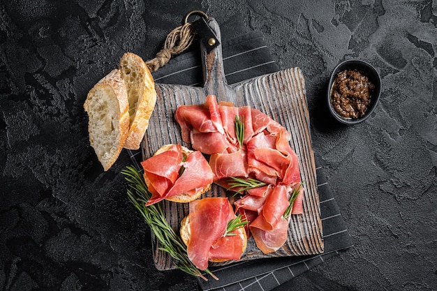 Toast aux tomates et séché Tranches de jambon serrano jamon prosciutto crudo parme sur planche de bois avec romarin Fond noir Vue de dessus