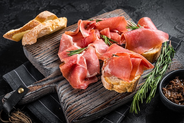 Toast aux tomates et tranches de jambon serrano, prosciutto crudo parme sur planche de bois au romarin. Fond noir. Vue de dessus.